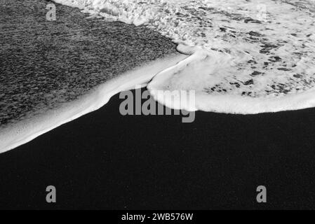 Spiaggia di sabbia nera per lo più sfocata con schiuma bianca di onde marine sullo sfondo Foto Stock