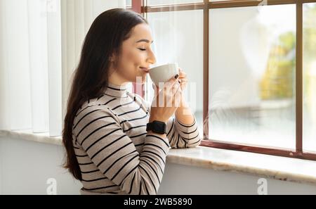 Una giovane donna soddisfatta con i suoi capelli eleganti in uno smartwatch e una camicia a righe assaggia il suo caffè Foto Stock