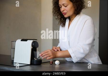 Donna di mezza età con i capelli ricci latinoamericani in accappatoio bianco che si sveglia la mattina e prepara un espresso fresco con la macchina da caffè a capsule Foto Stock