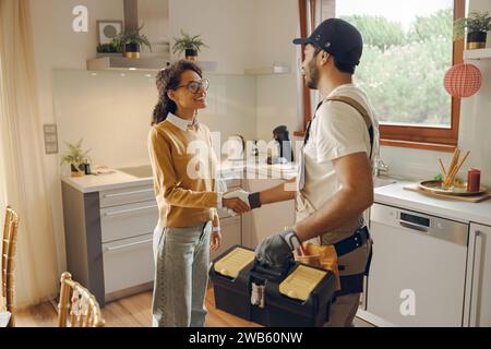 Riparatore professionista in uniforme che stringe la mano con una donna mentre si trova in cucina Foto Stock