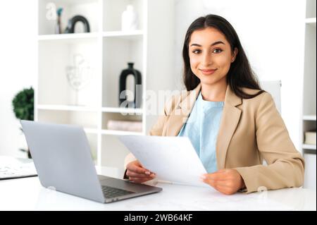 Donna d'affari indiana o araba bruna di successo sicura di sé in un elegante abito, ceo, dipendente aziendale, seduta a una scrivania nell'ufficio moderno, conserva i documenti, guarda la fotocamera e sorride Foto Stock