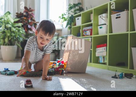 Un allegro ragazzo europeo gioca con le auto sul tappeto nella sua camera. Foto Stock