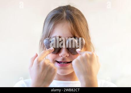 Primo piano felice gioioso che ride il bambino che chiude gli occhi davanti a due biscotti dolci a forma di cuore nelle sue mani, indulge. Bambina in posa su backgroun bianco Foto Stock