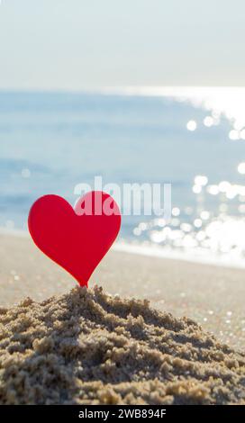 Bastoncino di plastica a forma di cuore rosso nella sabbia sulla spiaggia sabbiosa della riva del mare sullo sfondo delle onde del mare in un primo piano soleggiato giorno d'estate. Concetto amore relazione romanticismo amorosità amour St. San Valentino Foto Stock