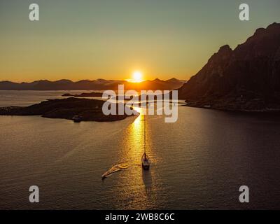 Una barca a vela sotto il sole di mezzanotte, dietro una grande collina a Lofoten, vicino a Henningsvaer, in lontananza alte montagne, ponte che collega Henningsvaer. Foto Stock