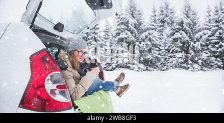 Donna con cappello di lana siede nel bagagliaio dell'auto e tiene una tazza di tè caldo in mano. Foto Stock