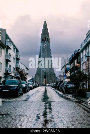 Vista della chiesa di hallgrimskirkja a Reykjavik, Islanda, situata alla fine di una strada fiancheggiata da edifici e auto parcheggiate, sotto un cielo nuvoloso. La Foto Stock