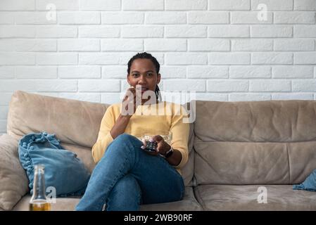 Ridere la giovane e graziosa donna afro-americana seduta sul divano, mangiare popcorn e guardare la TV a casa, tenere il telecomando e godersi un bel film Foto Stock