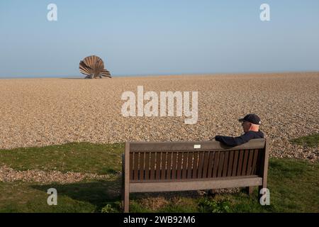 Uomo seduto sulla panchina con le conchiglie di capesante sulla spiaggia. Tra Aldeburgh e Thorpeness Suffolk, Inghilterra Foto Stock