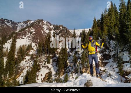 Un turista fa trekking in montagna in inverno. Un uomo barbuto con zaino e bastoncini da trekking indossa una giacca gialla. Stile di vita attivo Foto Stock