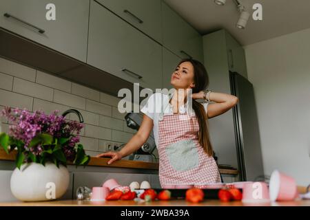 Felice cuoca in piedi nella sua nuova cucina sorridente, con grembiule e guanti da forno pronti per cuocere. Bella giovane razza mista Caucasi Foto Stock