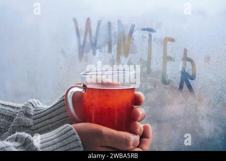 Le mani in un caldo maglione bianco tengono una tazza calda di tè vicino alla finestra in una giornata invernale. Stile di vita casalingo invernale Foto Stock