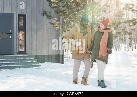 Felice giovane coppia interculturale in abbigliamento invernale che cammina lungo il sentiero della neve e parla o chiacchiera durante il fine settimana invernale Foto Stock