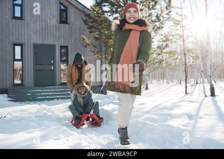 Giovane donna felice che tira la slitta con il suo simpatico figlio mentre un uomo afro-americano lo spinge indietro durante la passeggiata invernale Foto Stock