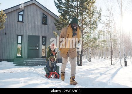 Felice giovane afro-americano che tira la slitta con un ragazzo carino mentre sua moglie spinge il figlio sulla schiena durante la passeggiata invernale in campagna Foto Stock