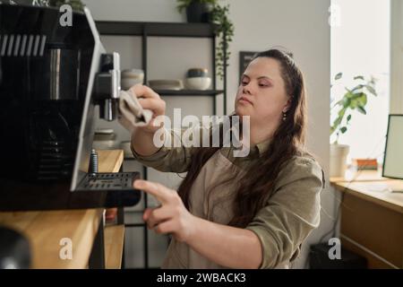 Giovane donna con disabilità mentale che pulisce i dettagli della macchina da caffè con la polvere umida prima di preparare il cappuccino per i clienti della caffetteria Foto Stock