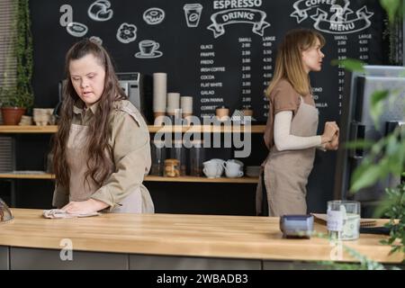 Giovane cameriera con disabilità che prepara il tavolo per gli ospiti del caffè al mattino e pulisce il tavolo con la polvere umida contro il collega Foto Stock