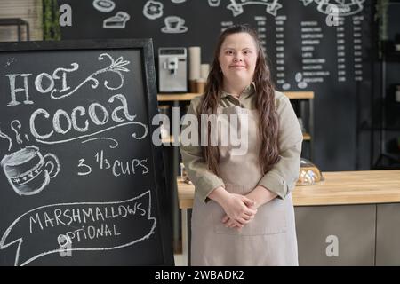 Cameriera sorridente con sindrome di Down che guarda la macchina fotografica mentre si trova accanto alla lavagna con un'offerta speciale del giorno scritta con gesso bianco Foto Stock