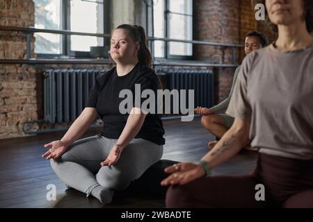 Giovane donna attiva con sindrome di Down seduta in posa di loto con gli occhi chiusi e meditando tra le altre persone in palestra Foto Stock
