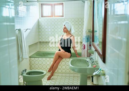 La donna si rilassa in un bagno in stile vintage, con accappatoio di seta, telo avvolto di turbante, serenità post-bagno. Siediti vicino alla vasca, guarda lontano, rifletti su se stessi Foto Stock