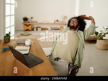 Giovane studente uomo pigro in abbigliamento casual che si allunga all'indietro mentre si siede sulla sedia di fronte al notebook e guarda le informazioni online Foto Stock