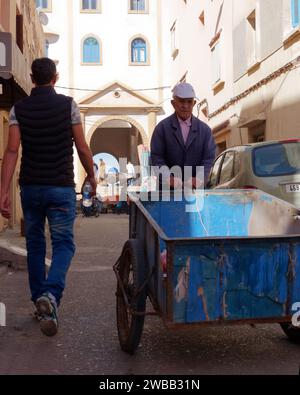 Un anziano in tuta in strada spinge un carro blu di legno utilizzato per trasportare bagagli e merci nella città di Essaouira, in Marocco. 9 gennaio 2024 Foto Stock