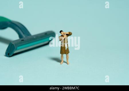 Fotografia di figure giocattolo in miniatura. Un uomo che indossa un accappatoio che si fa radere la barba accanto al rasoio. Isolato su sfondo verde. Foto immagine Foto Stock