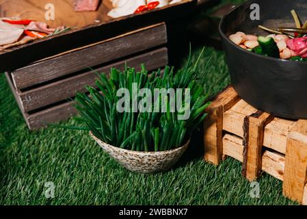 Un cesto pieno di erba cipollina fresca su una superficie di erba artificiale con casse rustiche di legno e una ciotola di verdure sullo sfondo. Foto Stock