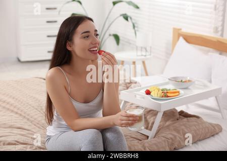 Una giovane donna felice che fa colazione vicino a un vassoio bianco a letto a casa Foto Stock