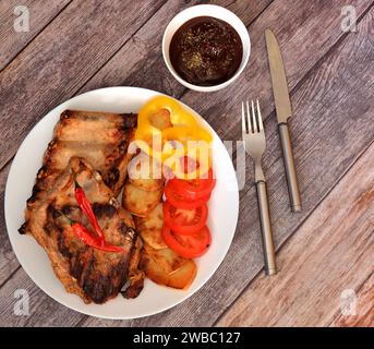 Costolette di maiale alla griglia con patate, pomodori, peperoni dolci e caldi su un tavolo di legno leggero, salsa e posate nelle vicinanze. Vista dall'alto, base piatta. Foto Stock