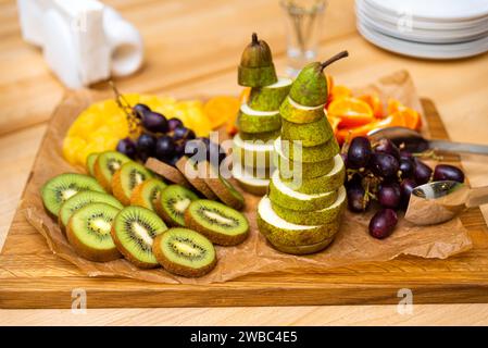 Una presentazione artistica di frutta a fette, tra cui una torre di fette di pera, kiwi e uva, esposta su una tavola di legno per un evento. Foto Stock
