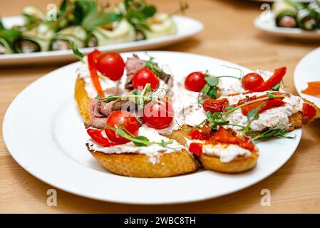 Deliziosa bruschetta condita con formaggio cremoso, succulente fette di manzo, pomodori ciliegini freschi e microgreen, disposta su un piatto bianco. Foto Stock