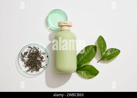 Scena di mockup per la pubblicità di cosmetici estratti dal tè verde. Una bottiglia verde senza marchio decorata su sfondo bianco con foglie di tè verde fresco, r Foto Stock