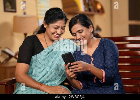 figlia sorridente con madre impegnata a usare il telefono cellulare a casa - concetto di condivisione sui social media, tecnologia e legame con la famiglia. Foto Stock