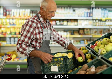 Uomo anziano del supermercato, dipendente di un negozio di alimentari. Mettere il cibo sugli scaffali del reparto verdure Foto Stock