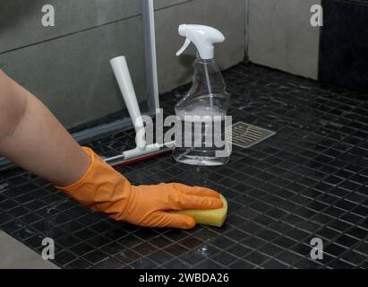 Primo piano della mano di una donna con guanti arancioni per pulire le piastrelle sporche del pavimento della doccia Foto Stock