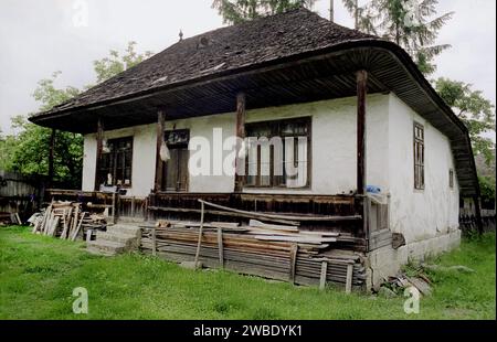 Contea di Vrancea, Romania, circa 1995. Una casa tradizionale con tetto in legno in cattive condizioni. Legno impilato nel cortile. Foto Stock