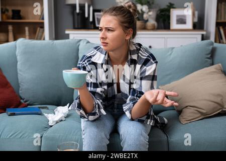 Giovane donna seduta a casa la mattina bevendo il suo primo caffè come routine quotidiana sentendosi sconvolta e irritata dal rumore forte del lavoro su strada sulla st Foto Stock