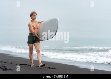Sportiva surfista in bikini top, pantaloncini corti e piedi nudi che cammina su una spiaggia di sabbia nera e che trasporta tavole da surf bianche sullo sfondo delle onde del mare Foto Stock