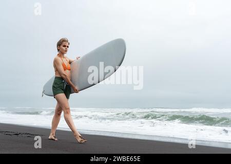 Surfista femminile, vestita con top in bikini, pantaloncini e piedi scalzi, cammina lungo la spiaggia di sabbia nera mentre trasporta tavole da surf bianche sullo sfondo delle onde dell'oceano Foto Stock