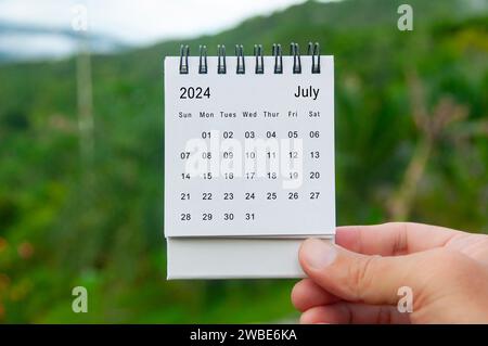 Mano che tiene il calendario bianco del 2024 luglio con sfondo naturale. Concetto di calendario e festività. Foto Stock