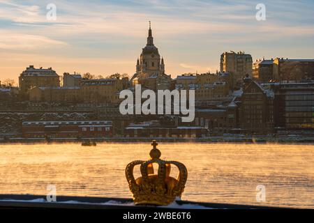 Simbolo della corona reale svedese, color oro, davanti allo skyline di Stoccolma in inverno, con la vecchia chiesa di Katarina sullo sfondo. Foto Stock
