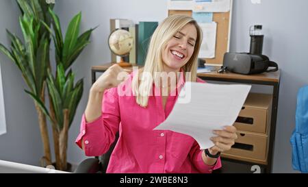 Una donna bionda allegra che indossa una camicia rosa esulta mentre legge un giornale in un moderno ambiente da ufficio. Foto Stock