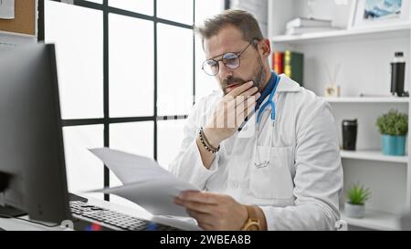 Un uomo pensivo di mezza età con i capelli grigi, che indossa uno stetoscopio, esamina i documenti in uno studio ospedaliero. Foto Stock