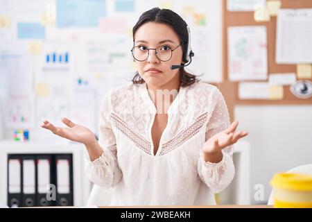 Giovane donna ispanica che lavora in ufficio, indossa cuffie e occhiali che urla e urla a tutto volume con la mano sulla bocca. concetto di comunicazione. Foto Stock
