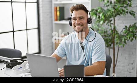 Un giovane ispanico sorridente con la barba che indossa un auricolare che lavora su un computer portatile in un ufficio moderno. Foto Stock