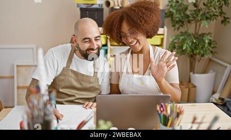 Due artisti sorridenti, un uomo e una donna, che conducono con sicurezza una videochiamata in uno studio d'arte Foto Stock