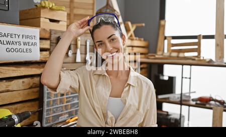 Bellissima giovane donna ispanica, un falegname sorridente, orgogliosa della sua officina di falegnameria, che indossa occhiali di sicurezza Foto Stock