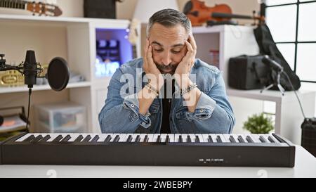 Uomo ispanico pensivo con barba grigia in studio con tastiera e microfono, che denota un'atmosfera musicale creativa. Foto Stock