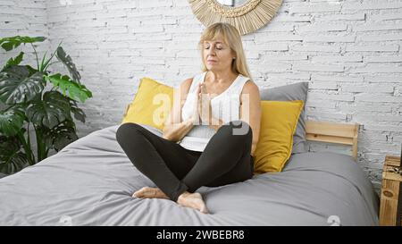 Sveglia e concentrata, la donna bionda di mezza età si diverte ad allenarsi con yoga mattutino seduto sul letto, abbracciando calma ed equilibrio nella sua camera da letto Foto Stock